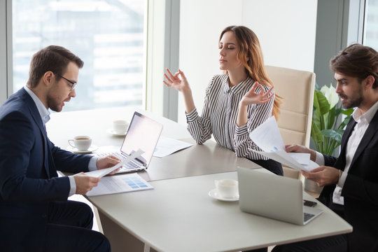 Female worker meditating at workplace staying calm while male colleagues stress about contract disagreement or falling rates and bad statistics, peaceful woman practice yoga at stressful meeting