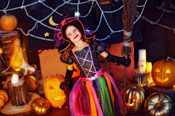 Happy Halloween. Cute little child girl in a witch costume with magic broom celebrates at home in an interior with pumpkins and cardboard magic house on the background.