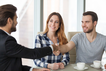 Excited millennial husband shaking hand of professional realtor or agent closing deal, satisfied couple handshaking consultant or banker happy to make agreement or greeting visiting at his office