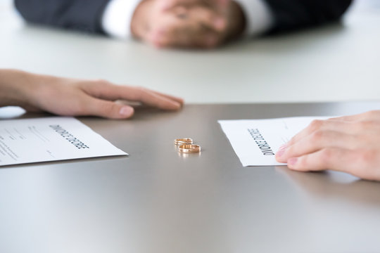 Close up of marriage rings lying on table along with divorce decrees and couple hands, husband and wife sign paper separating or breaking up officially, spouses end relationships at lawyer office