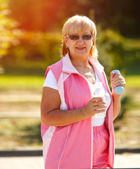 Pensioner woman sport. Adult woman is engaged in outdoor sports. Sport woman holding dumbbells and bottle of water. Active lifestyle. Happy woman.