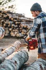 Bearded brutal lumberjack wearing plaid shirt sawing tree with chainsaw for work on sawmill....