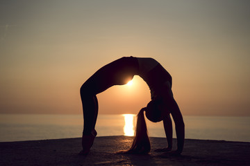 Silhouette of young gymnast doing a wheel pose on the beach at sunset. Woman practicing yoga or...
