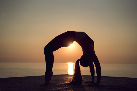 Silhouette of young gymnast doing a wheel pose on the beach at sunset. Woman practicing yoga or pilates on the pier. Bridge Pose, Urdhva Dhanurasana (Upward Bow), Chakrasana (Wheel). Side view.