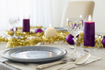 Close-up of a decorated dining table with burning candles, sterling cutlery, crystal glasses, colorful christmas balls and gold tinsel