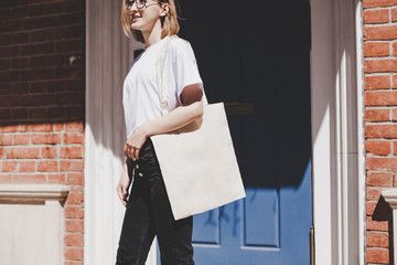Young hipster smiling girl with white blank cotton shopper paper bag wearing white t-shirt and...