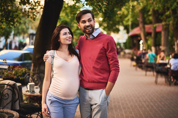 Fototapeta premium Loving couple. Portrait of beautiful young lady looking at husband with happy expression while he embracing her. They standing on sidewalk near outdoor cafe