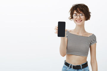 Look what I got. Portrait of pleased excited cute woman in round glasses and cropped top, smiling broadly while showing smartphone, talking about new app she downloaded recently