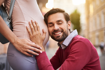 I can hear you. Portrait of happy father with closed eyes putting ear to lady tummy and smiling