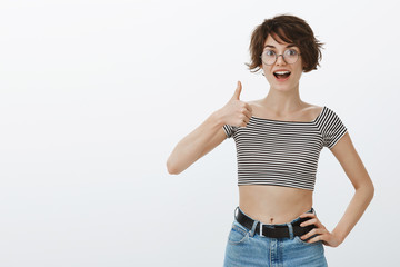 Studio shot of active joyful young woman in round glasses and striped tank top, holding hand on hip and showing thumb up while smiling broadly, agreeing or giving approval, liking new idea