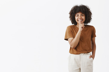 Happy carefree african-american cute teenage girl with afro hairstyle in stylish brown t-shirt and white pants holding hand in pocket casually smiling, showing shush sign with index finger over mouth