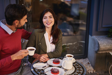 I adore you. Young bearded man with cup of tea hugging charming lady while she is looking at camera with smile. They sitting at the table with raspberry cakes and tea-set