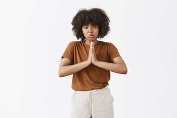 Studio shot of gloomy upset good-looking dark-skinned teenage woman with afro hairstyle pursing lips in sad smile frowning and holding hands in pray while asking help or favor over gray wall