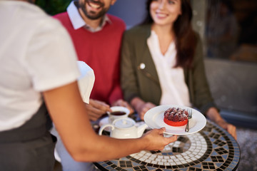 Obraz na płótnie Canvas Bon appetit. Female arm of cafe worker holding plate with raspberry cake. Smiling young lady and man sitting at the table on blurred background