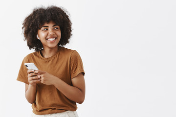 Studio shot of carefree friendly and creative stylish african american teenage girl in brown t-shirt turning right with broad satisfied smile wearing wireless earphones and holding smartphone