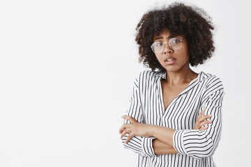 Waist-up shot of silly and timid worried dark-skinned female model with curly hair in glasses and office striped blouse holding hands on chest frowning form empathy and anxiety over gray wall