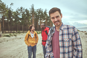 Obraz premium Rejoicing with friends. Waist up portrait of happy man standing and smiling while two of his friends joking on background