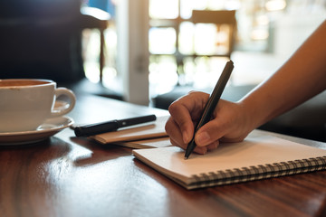 Closeup image of a hand writing down on a white blank notebook with coffee cup on wooden table