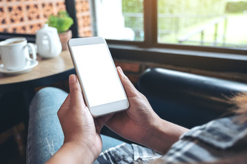 Mockup image of a woman's hands holding and using a white mobile phone with blank screen with coffee cup on table in modern cafe
