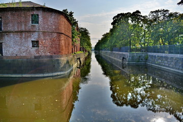 old ages medieval red brick fortress wall with overgrown sprouts of trees. Moat with water around it fortified stone on a Sunny summer day