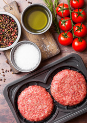 Plastic tray with raw minced homemade beef burgers with spices and herbs. Top view. On top of wooden kitchen table background with tomatoes salt and pepper.