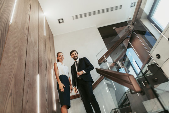 Low Angle Portrait Of Cheerful Businesswoman Talking With Beaming Bearded Male Colleague While Standing On Stairwell Landing