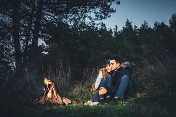 Pretty couple relaxing near bonfire in the forest at evening time