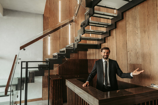 Portrait Of Happy Unshaven Male Employee Gesticulating Hand While Locating At Counter In Hotel