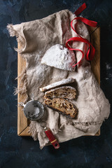 Sliced Christmas cake, traditional German festive baking. Wholegrain stollen with raisins and sugar powder on linen napkin, wooden tray, sieve, red ribbon over dark blue background. Top view, space