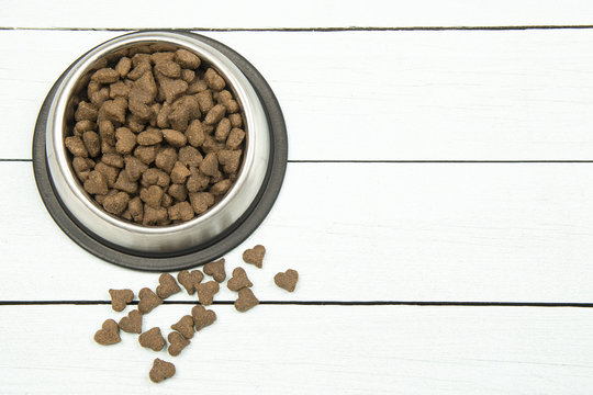 Metal round feeding bowl with dark brown heart shaped pet kibble on a white wooden background with space for copy