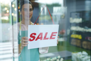 Waist up portrait of cheerful female shopkeeper hanging red SALE sign on glass door or window