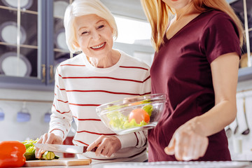 Cooking salad. Positive emotional senior woman leaning to the table with the knife in her hands while looking at the screen of a modern tablet
