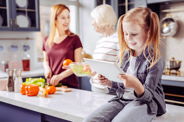 Child with tablet. Cheerful relaxed little girl looking at the screen of a modern tablet while her granny and mother cooking meals in the kitchen