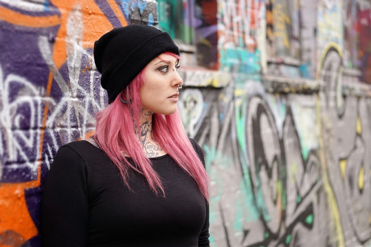 young alternative woman with pink hair piercings and tattoos leaning against graffiti wall                              