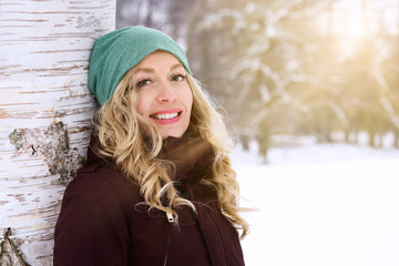 smiling woman leaning against birch tree enjoying sunny day in snow covered winter landscape, with sun flare light leak