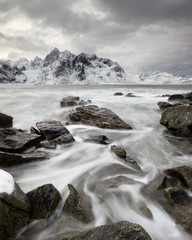 Fototapeta na wymiar Coastal landscape in winter with water movement between big stones, in the background a mountain range with snow, high contrast sky - Location: Norway, Lofoten