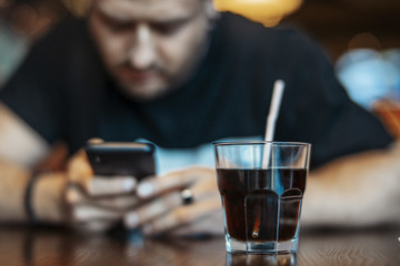 Young attractive man looking at mobile phone screen and drinking soda.