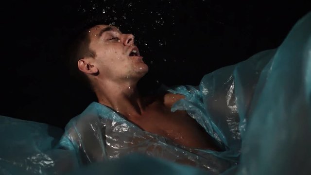 Sporty positive active guy with cute big blue eyes and stylish haircut with no clothes put on is making swimming movements in blue cellophane film imitating water waves and spits out real water.