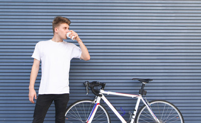 Young student wearing a white T-shirt stands by a bicycle on the background of a gray wall, drinks...