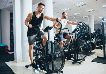 Young men with muscular body using air bike for cardio workout at cross training gym.