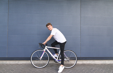 Happy young man sitting on a white bike against the wall, looking at the camera and smiling. Guy...