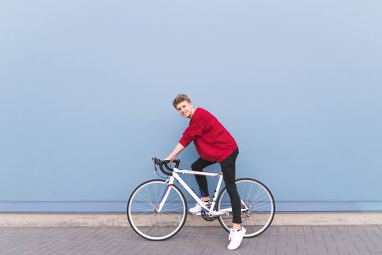 Happy young man in a red sweatshirt sitting on a white bike against a blue wall background. Stylish smiling cyclist on a blue background. Copyspace