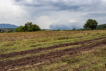 Muddy wheel tracks in a field at the foot of a Rhodope Mountains hill under dramatic stormy sky in Bulgaria