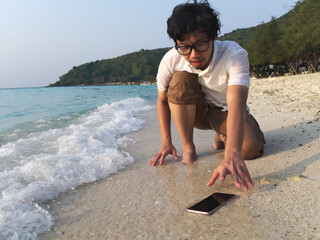 Shocked Asian man dropping mobile smart phone on tropical sandy beach of sea. Accident and insurance electronic equipment concept
