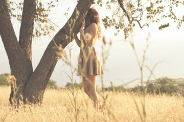 girl on nature, young woman walking in summer field at sunset hiding behind a tree