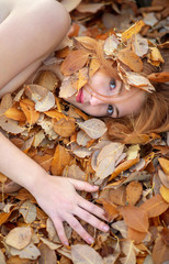 Schönes sexy reizendes junges rothaariges Mädchen, das auf goldenem Herbstlaub liegt, bedeckt mit gelben herbstlichen Blättern, im Park, mit freundlichem Lächeln im Gesicht.