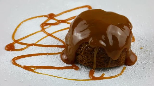 Sticky sponge pudding rotating close up on a white sugar dusted plate drizzled with toffee sauce