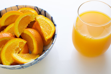 Freshly squeezed orange juice in a glass next to a bowl of orange slices and two oranges just picked off the tree on a white table for breakfast.