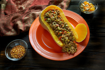 Fall colors with Butternut Squash Boat filled with ground beef with sliced olives on a red plate with some red pepper flakes on the side for a spicy kick on a wooden kitchen table.