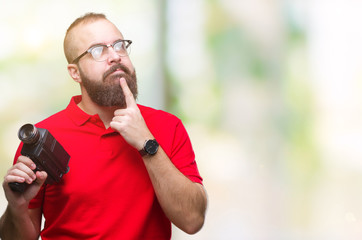 Young caucasian hipster man filmming using retro video camera over isolated background serious face thinking about question, very confused idea